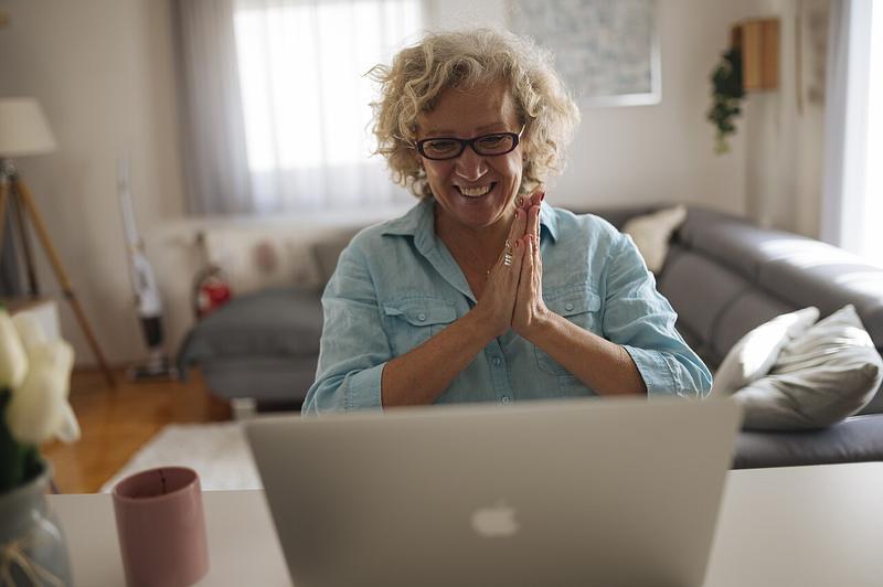 A smiling older woman on a video call at home, hands pressed together