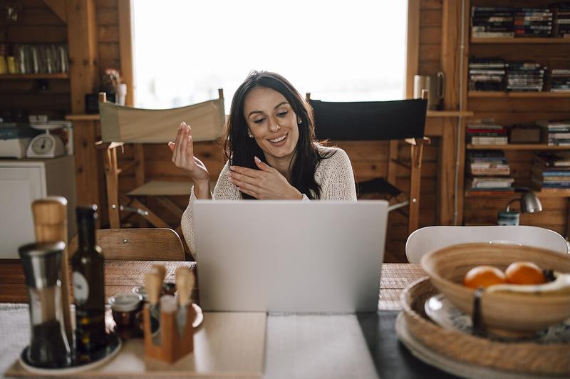 A smiling woman talking on a video call on her laptop at a kitchen table